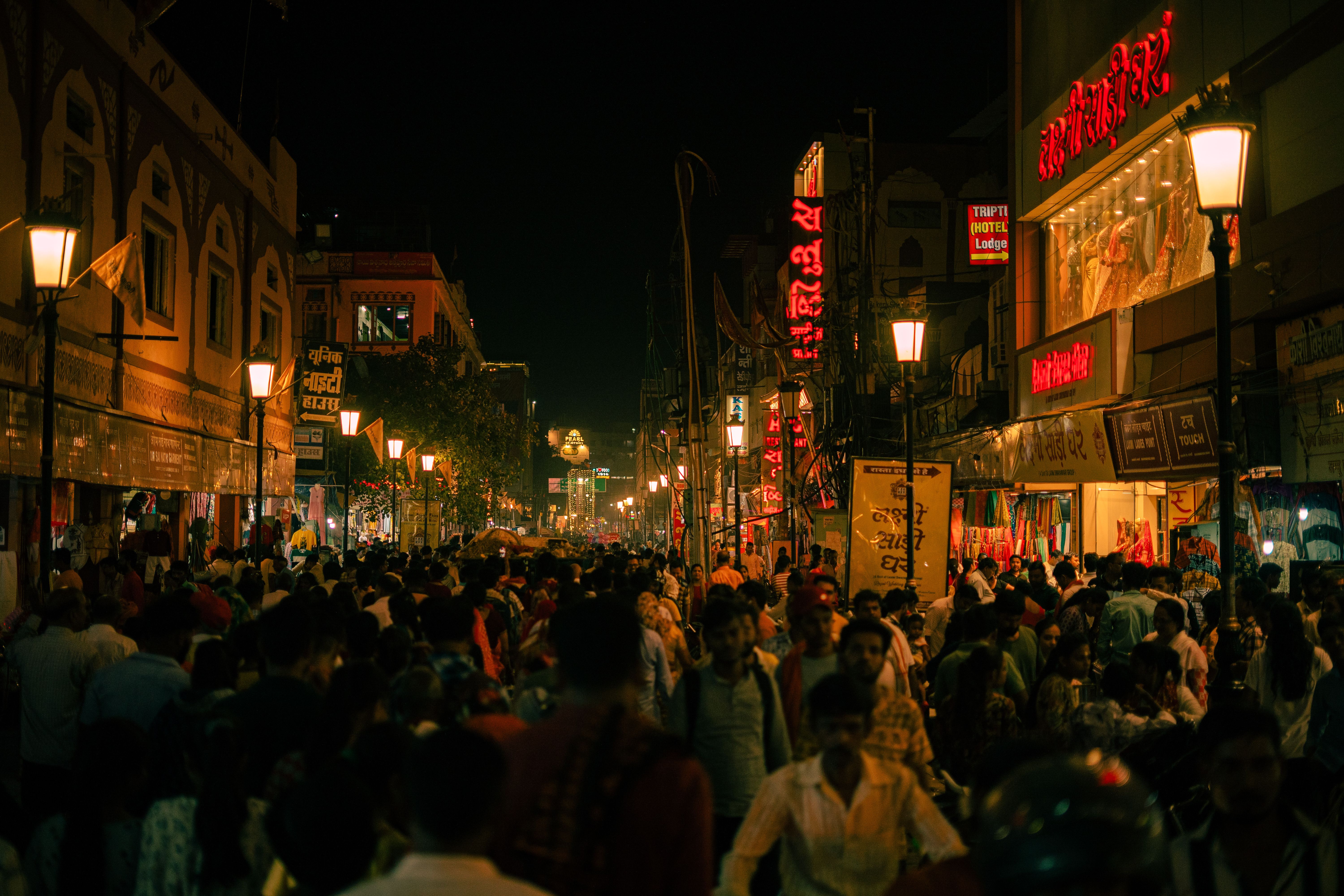 A crowded Indian market street at night lit by lamps and neon shop signs, filled with shoppers