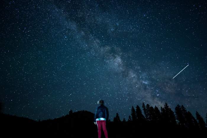 Silhouetted person standing alone looking up at a starry night sky with the Milky Way and a shooting star