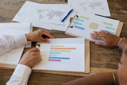 Two people at a table pointing at and discussing printed bar charts and data sheets together