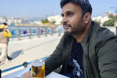 Rahul Phukan sitting at a waterfront cafe table with a beer, looking out over a harbour